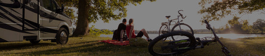 Two people sits with bikes and their Tiffin motorhome looking out over a lake during a sunset